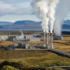 Elektrownia geotermalna z wysokimi kominami wydzielającymi parę, otoczona surowym krajobrazem i górami w tle.