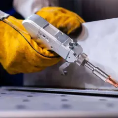 Ręce w żółtych rękawicach trzymają laserową spawarkę, która precyzyjnie łączy metalowe elementy, minimalizując wady spawalnicze.