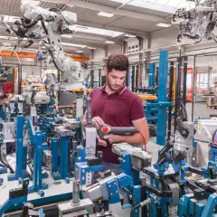 Dwóch pracowników w hali produkcyjnej, otoczonych robotami przemysłowymi i skomplikowanymi maszynami. Jeden z nich, w białym kasku, trzyma tablet. Drugi, z narzędziem w ręku, pracuje przy linii montażowej. Widać wyraźne braki kadrowe, bo to oni muszą o...