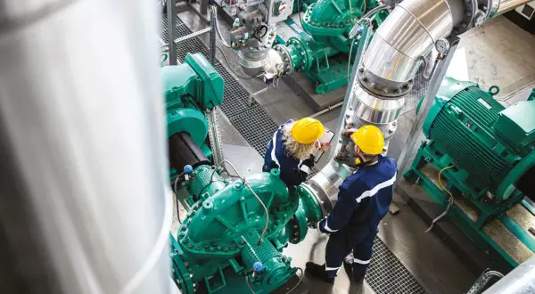 Dwóch inżynierów w żółtych kaskach sprawdza działanie pomp przemysłowych, dbając o oszczędzanie energii.