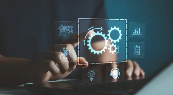 Dłoń dotyka interfejsu z ikonami symbolizującymi złożony system informatyczny: trybiki, chip, wykres, lista zadań, żarówka i słuchawki.