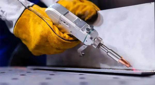 Ręce w żółtych rękawicach trzymają laserową spawarkę, która precyzyjnie łączy metalowe elementy, minimalizując wady spawalnicze.