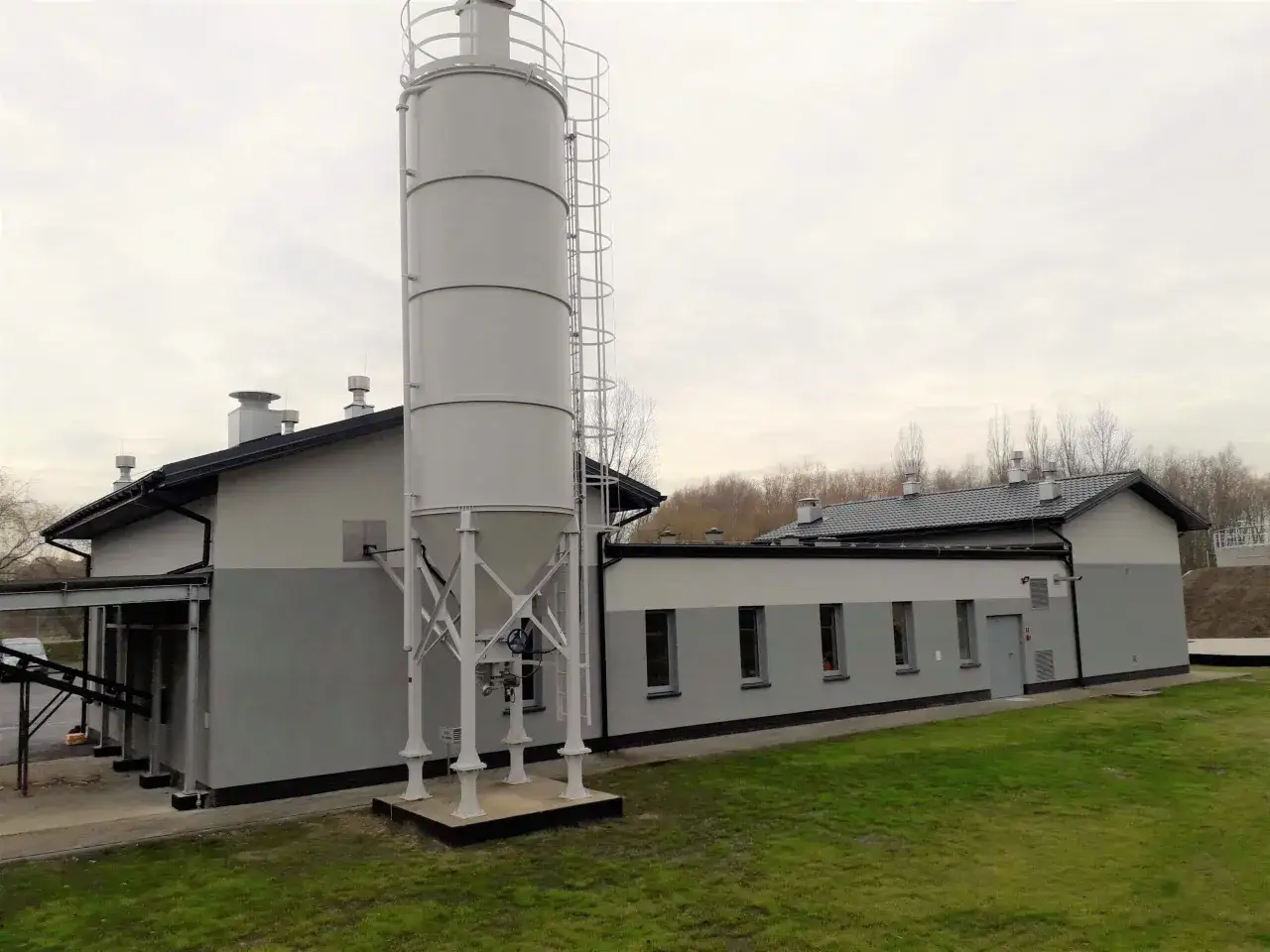 Wysoki, biały silos stoi obok szarego budynku przemysłowego. To właśnie co to jest silos – konstrukcja do przechowywania materiałów sypkich.