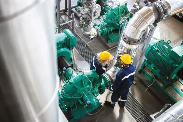 Dwóch inżynierów w żółtych kaskach sprawdza działanie pomp przemysłowych, dbając o oszczędzanie energii.