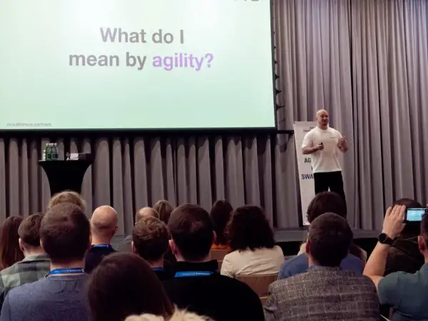 Prelegent wyjaśnia, co to agile, podczas konferencji. Na ekranie pytanie: "What do I mean by agility?".