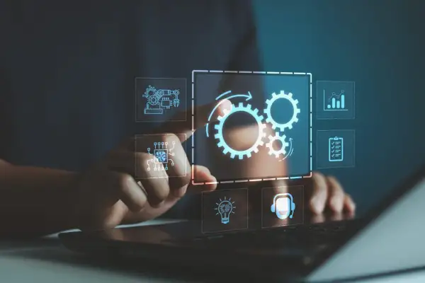 Dłoń dotyka interfejsu z ikonami symbolizującymi złożony system informatyczny: trybiki, chip, wykres, lista zadań, żarówka i słuchawki.