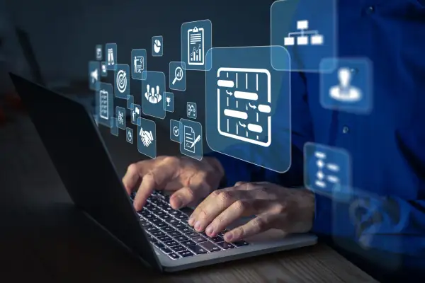 Dłonie pracują na laptopie, nad którym unoszą się ikony symbolizujące integrację systemów, zarządzanie projektami i współpracę.